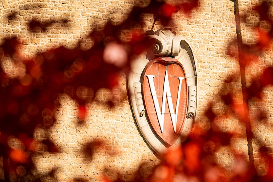 A W crest emblem on the side of the UW Field House is pictured through blurred autumn leaves in the foreground.