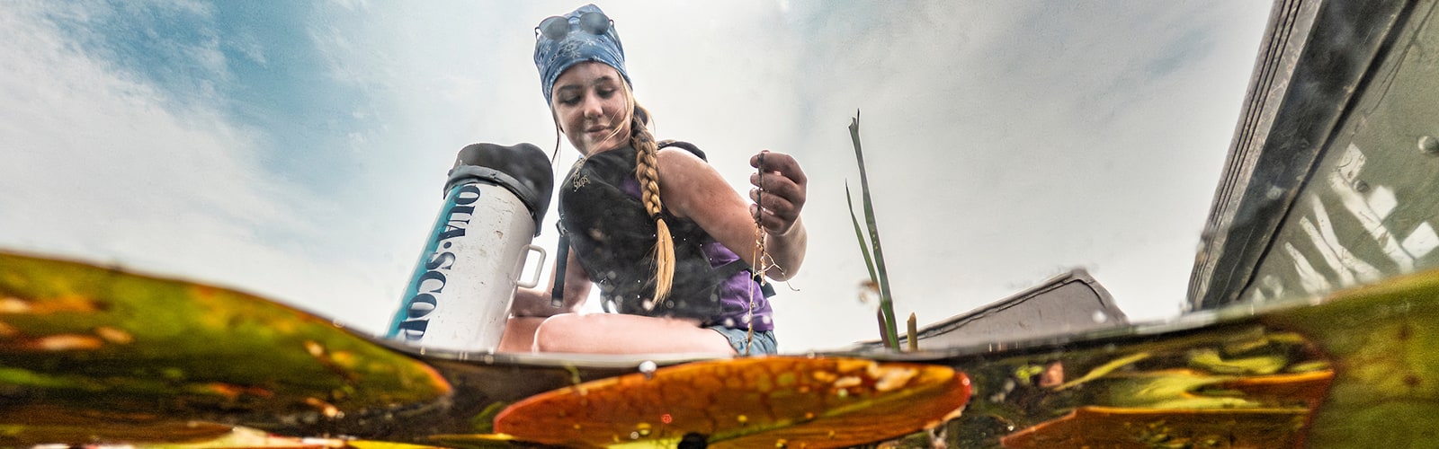 Photo taken at the waterline to show lilypads growing underwater and also a female student wearing a life vest sitting in a canoe, taking a sample of wild rice from the lake.