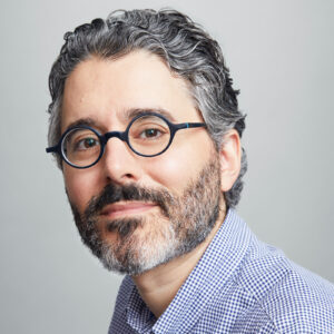 Headshot of Michael Barbaro, wearing round glasses and a blue checkered shirt.
